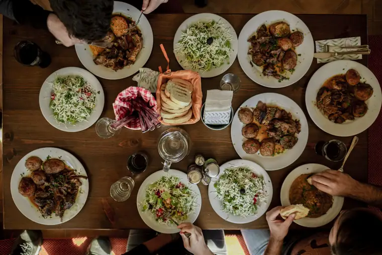 A long table seen from above, covered in shared plates, glasses and half a dozen reaching hands.